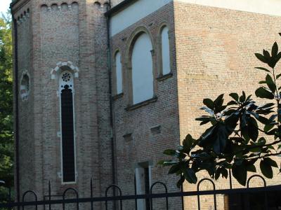 A brick building with a tower and arched windows, viewed from behind a black metal fence with green foliage in the foreground.