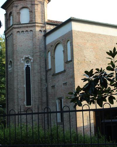 A brick building with a tower and arched windows, viewed from behind a black metal fence with green foliage in the foreground.