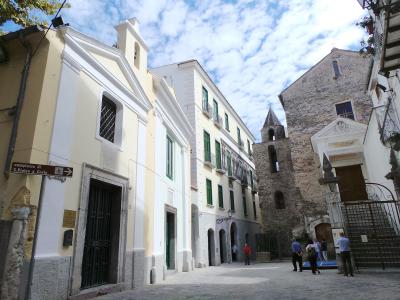 A street scene in Salerno, Italy, showing the pale yellow facade of the Chiesa di San Pietro a Corte on the left with a directional sign reading "complesso di S. Pietro a Corte," alongside adjacent buildings and a stone tower under a cloudy sky.