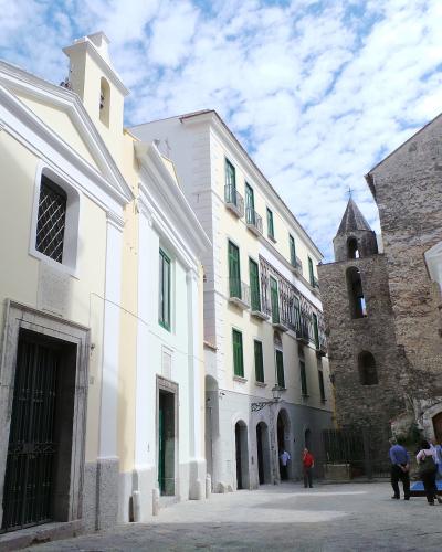 A street scene in Salerno, Italy, showing the pale yellow facade of the Chiesa di San Pietro a Corte on the left with a directional sign reading "complesso di S. Pietro a Corte," alongside adjacent buildings and a stone tower under a cloudy sky.
