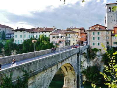 A stone arch bridge with pedestrians and a car crossing it, set against a backdrop of historic Italian-style buildings and a prominent bell tower under a cloudy sky.