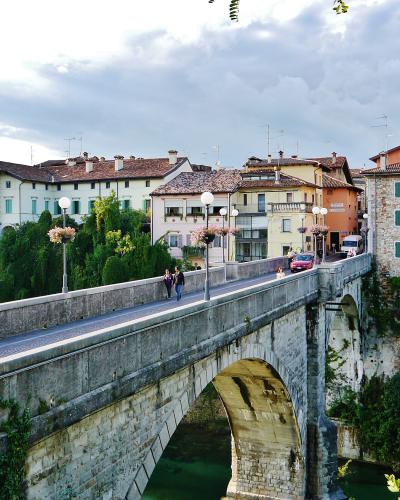 A stone arch bridge with pedestrians and a car crossing it, set against a backdrop of historic Italian-style buildings and a prominent bell tower under a cloudy sky.