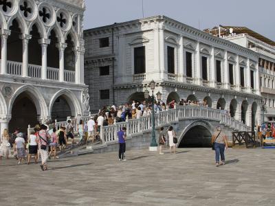 A stone bridge with a single arch and balustrades, crowded with tourists, situated between the Doge's Palace on the left and the Procuratie Nuove on the right, overlooking a canal in Venice.