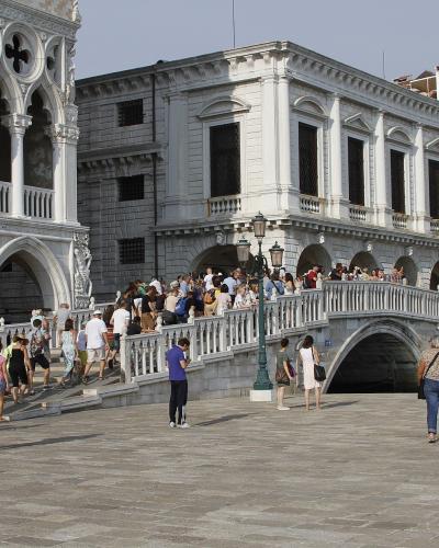 A stone bridge with a single arch and balustrades, crowded with tourists, situated between the Doge's Palace on the left and the Procuratie Nuove on the right, overlooking a canal in Venice.