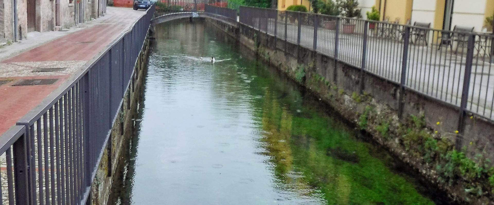 A narrow, man-made canal or river channel flows through a small Italian town, flanked by stone and stucco buildings, with a metal railing along the left bank and a yellow building on the right; a sign reading “OSTERIA” is visible in the background