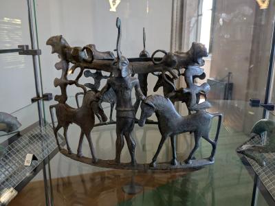 A bronze Etruscan-style fibula (brooch) depicting a central human figure flanked by horses and other animals, displayed inside a glass museum case with a small white label marked "10" visible on the left.