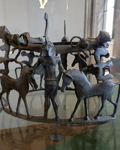 A bronze Etruscan-style fibula (brooch) depicting a central human figure flanked by horses and other animals, displayed inside a glass museum case with a small white label marked "10" visible on the left.