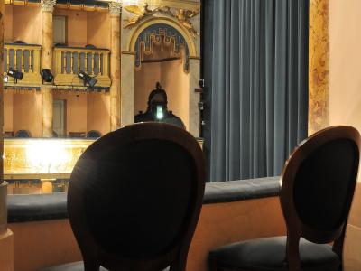 The image shows the interior of a historic theater box, featuring two dark wooden chairs with blue upholstery in the foreground, looking out over a stage area with a large blue curtain, ornate gold and marble-style architectural details, statues o