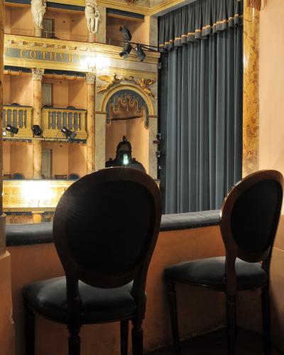 The image shows the interior of a historic theater box, featuring two dark wooden chairs with blue upholstery in the foreground, looking out over a stage area with a large blue curtain, ornate gold and marble-style architectural details, statues o