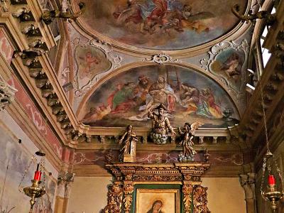 The image shows the ornate interior of a church, featuring a gilded altar with a painting of the Madonna and Child, flanked by candles and hanging lamps, with elaborate frescoes and stucco work on the vaulted ceiling.