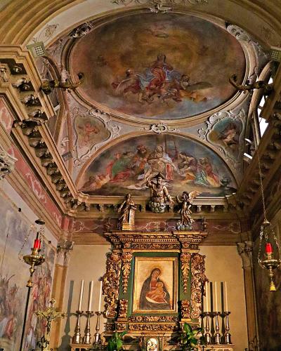 The image shows the ornate interior of a church, featuring a gilded altar with a painting of the Madonna and Child, flanked by candles and hanging lamps, with elaborate frescoes and stucco work on the vaulted ceiling.