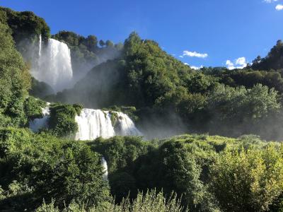 A tiered waterfall cascading down a lush, green, forested hillside under a bright blue sky, with visible mist rising from the falls.