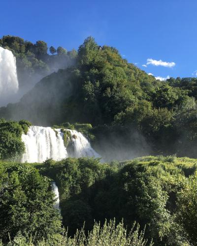 A tiered waterfall cascading down a lush, green, forested hillside under a bright blue sky, with visible mist rising from the falls.