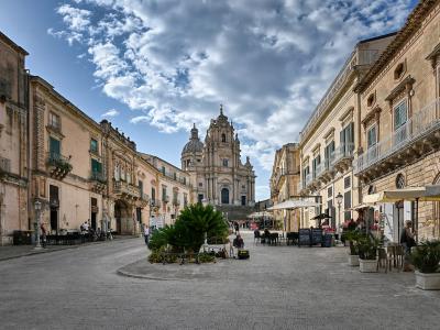 A wide, paved pedestrian plaza in a historic Italian town, flanked by multi-story buildings with balconies and shuttered windows, leading toward a large, ornate Baroque cathedral with a central dome and twin bell towers under a partly cloudy sky.