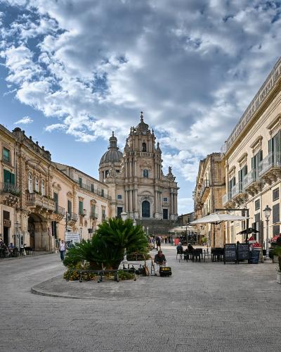 A wide, paved pedestrian plaza in a historic Italian town, flanked by multi-story buildings with balconies and shuttered windows, leading toward a large, ornate Baroque cathedral with a central dome and twin bell towers under a partly cloudy sky.
