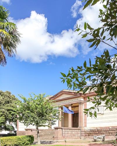 A low-angle exterior shot of a neoclassical building with a pediment and columns, partially framed by a palm tree on the left and leafy branches on the right; visible text on the facade reads “MUSEO NAZIONALE G.A. SANNA” beneath the pediment, with
