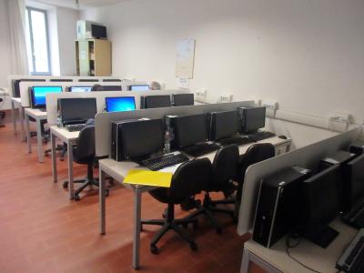 A computer lab with rows of desktop computers on desks, black office chairs, and terracotta-colored tiled flooring.