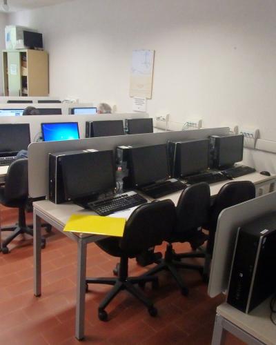 A computer lab with rows of desktop computers on desks, black office chairs, and terracotta-colored tiled flooring.