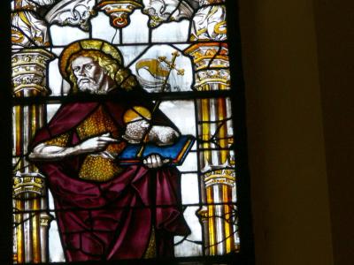 A close-up photograph of a stained-glass window depicting a bearded male saint with a halo, wearing purple and gold robes, holding a book and a staff with a cross, flanked by ornate columns and angelic figures above.