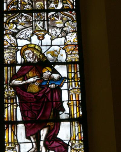 A close-up photograph of a stained-glass window depicting a bearded male saint with a halo, wearing purple and gold robes, holding a book and a staff with a cross, flanked by ornate columns and angelic figures above.