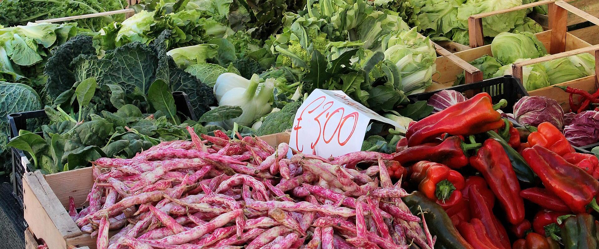 A close-up view of a vegetable stall at an outdoor market, featuring crates of fresh produce including pink-speckled beans, red peppers, leafy greens, and eggplants, with a handwritten price sign showing "€ 3.00".