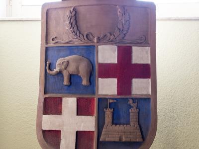 A painted wooden relief of the coat of arms of Catania, featuring an elephant, a castle, and crosses, displayed on an easel indoors.