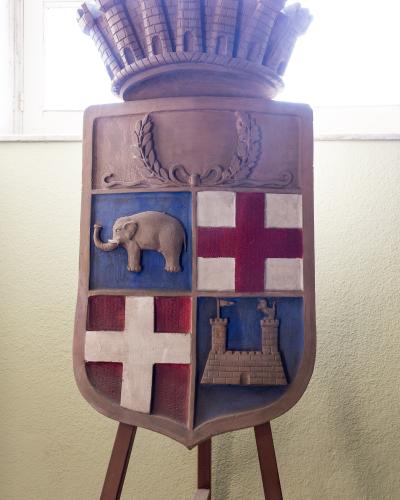 A painted wooden relief of the coat of arms of Catania, featuring an elephant, a castle, and crosses, displayed on an easel indoors.