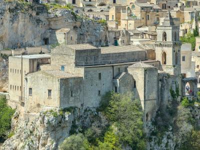 A view of the ancient rock-hewn church complex of San Pietro Barisano, perched on a limestone cliff edge with visitors visible on walkways and terraces, set against the backdrop of Matera’s historic Sassi district.