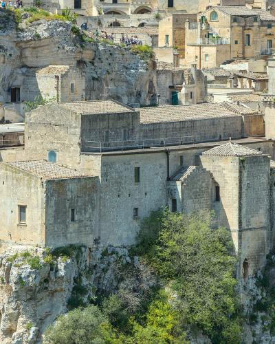 A view of the ancient rock-hewn church complex of San Pietro Barisano, perched on a limestone cliff edge with visitors visible on walkways and terraces, set against the backdrop of Matera’s historic Sassi district.