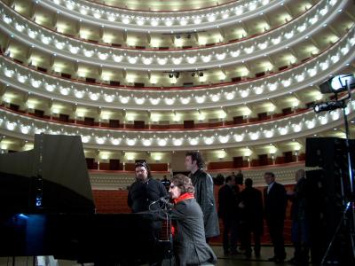 The image shows the interior of a multi-tiered opera house or theater with curved balconies, red seating, and a stage where a woman is singing at a piano with other people present.