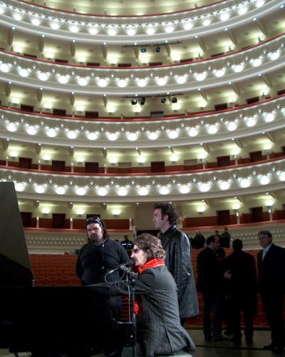 The image shows the interior of a multi-tiered opera house or theater with curved balconies, red seating, and a stage where a woman is singing at a piano with other people present.