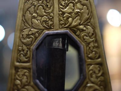 A close-up of an ornate, gilded wooden or plaster architectural element with baroque-style acanthus leaf carvings and a central octagonal inset containing a dark vertical object, possibly a candlestick or part of a liturgical stand, with blurred b