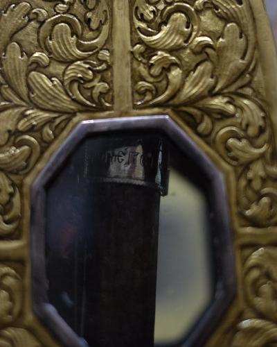 A close-up of an ornate, gilded wooden or plaster architectural element with baroque-style acanthus leaf carvings and a central octagonal inset containing a dark vertical object, possibly a candlestick or part of a liturgical stand, with blurred b