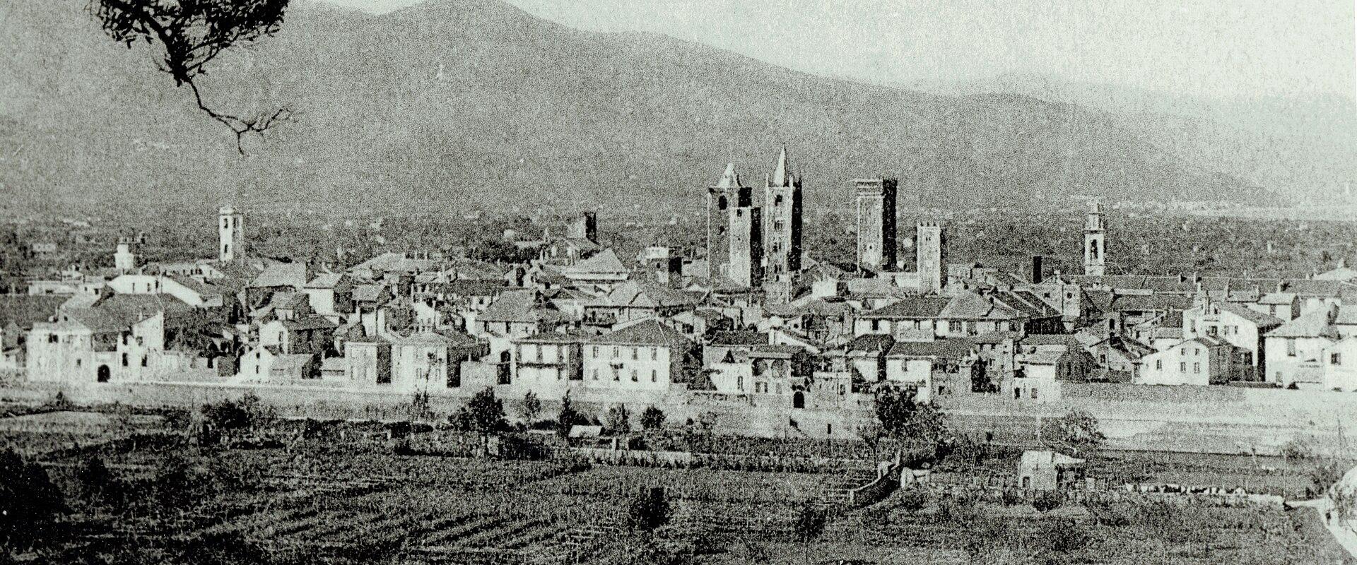A black-and-white historical photograph showing a walled Italian town with multiple prominent medieval towers and bell towers, viewed from an elevated position over cultivated fields, with mountains in the background; no visible signage or text.