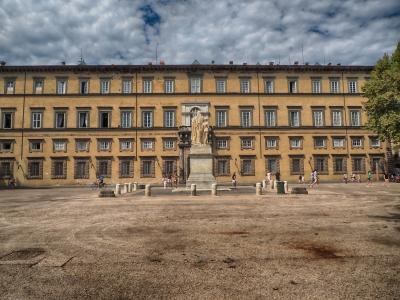 A wide, gravel-covered public square featuring a central statue on a pedestal in front of a large, three-story yellow palace with numerous windows and classical architecture.
