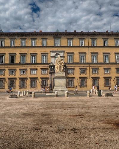 A wide, gravel-covered public square featuring a central statue on a pedestal in front of a large, three-story yellow palace with numerous windows and classical architecture.