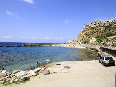 A sunny coastal scene showing a sandy beach with people swimming and sunbathing, a stone breakwater, a promenade with a parked silver SUV, and a historic town built on a steep cliff overlooking the sea.