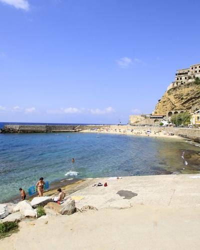 A sunny coastal scene showing a sandy beach with people swimming and sunbathing, a stone breakwater, a promenade with a parked silver SUV, and a historic town built on a steep cliff overlooking the sea.
