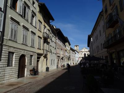 A sunlit, narrow European street paved with cobblestones, flanked by multi-story buildings with shuttered windows and balconies, leading toward a church facade in the distance under a clear blue sky.