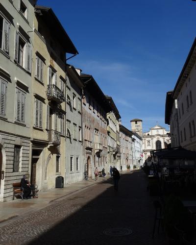 A sunlit, narrow European street paved with cobblestones, flanked by multi-story buildings with shuttered windows and balconies, leading toward a church facade in the distance under a clear blue sky.