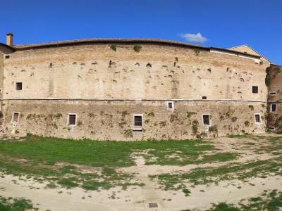 A wide-angle photograph of a large, curved brick and stone fortress with rounded bastions on either end, situated on a grassy and dirt-covered ground under a blue sky.