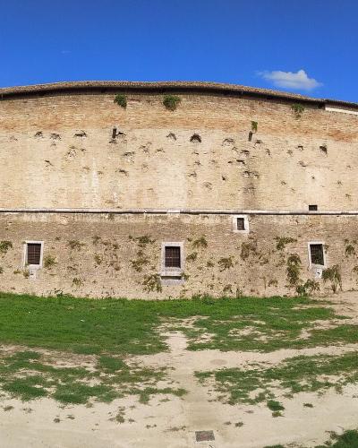 A wide-angle photograph of a large, curved brick and stone fortress with rounded bastions on either end, situated on a grassy and dirt-covered ground under a blue sky.