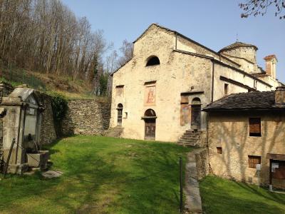 The image shows the exterior of the Church of San Costanzo al Monte, a historic stone church with a frescoed facade and a small bell tower, situated on a grassy slope with a stone wall and fountain in the foreground.