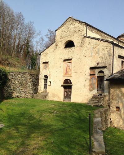 The image shows the exterior of the Church of San Costanzo al Monte, a historic stone church with a frescoed facade and a small bell tower, situated on a grassy slope with a stone wall and fountain in the foreground.