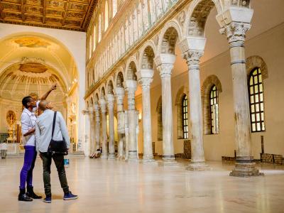 The image shows the interior nave of a basilica with a colonnade of tall, slender columns supporting an arcade on the right, a high clerestory level with mosaics above, and a coffered wooden ceiling; two visitors are looking up toward the apse are