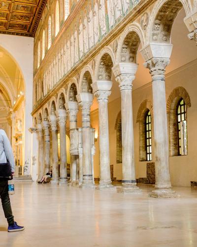 The image shows the interior nave of a basilica with a colonnade of tall, slender columns supporting an arcade on the right, a high clerestory level with mosaics above, and a coffered wooden ceiling; two visitors are looking up toward the apse are