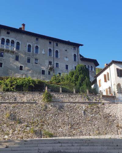 A low-angle view of a large, multi-story stone palace with arched windows and a balcony, situated atop a high, sloping stone retaining wall under a clear blue sky.