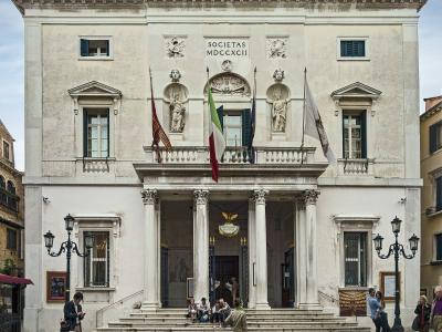 A frontal view of the neoclassical facade of Teatro La Fenice in Venice, featuring four Corinthian columns, a central balcony with statues, and the inscription "SOCIETAS MDCCXCII" above the entrance.