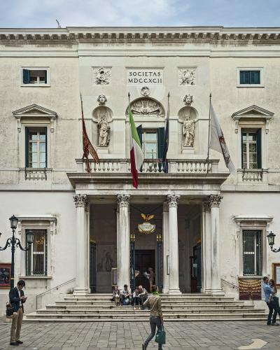 A frontal view of the neoclassical facade of Teatro La Fenice in Venice, featuring four Corinthian columns, a central balcony with statues, and the inscription "SOCIETAS MDCCXCII" above the entrance.