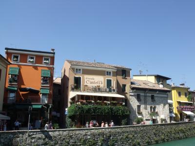 A street-level view of colorful buildings along a waterfront promenade in Sirmione, featuring the "Piccolo Castello" restaurant with outdoor seating and people walking nearby.